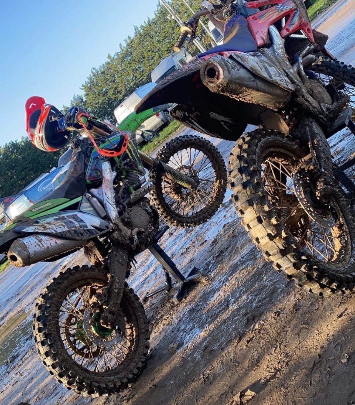 Two mud-covered motocross bikes on stands in a sunny paddock