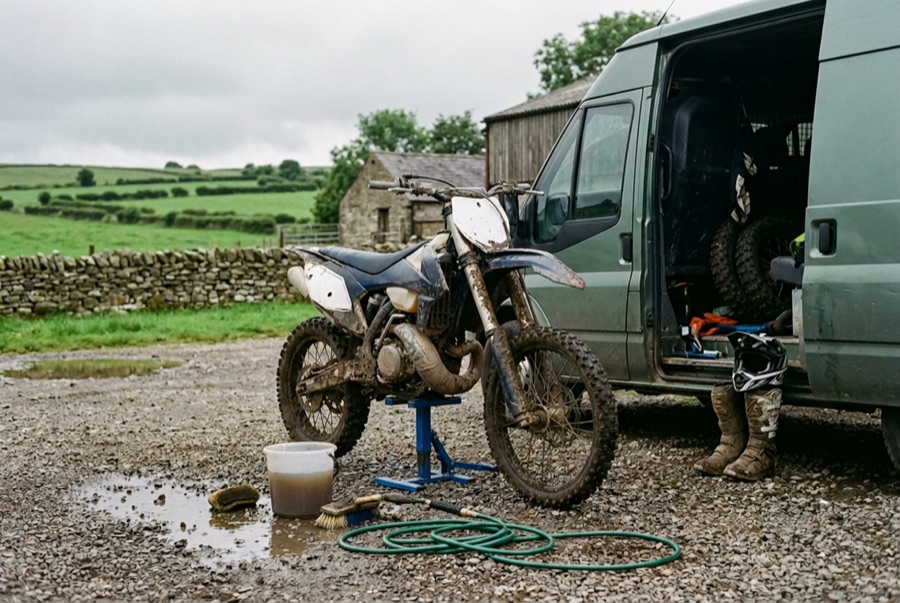 A muddy dirt bike on a stand beside a van, with a bucket, brush and hose ready for a wash